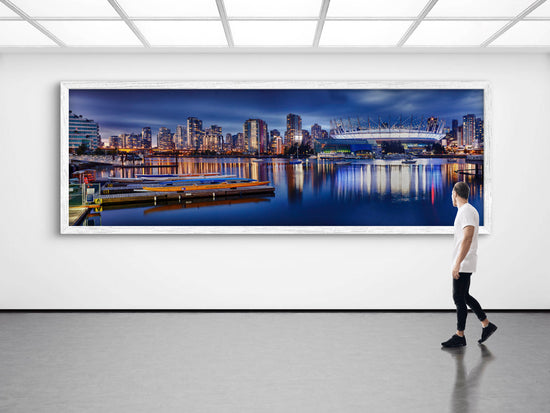Man walking past a large framed panoramic cityscape of a waterfront at night.