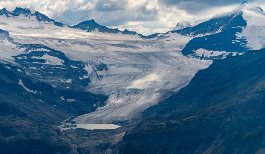 A dramatic mountain landscape with a large glacier flowing down a valley, surrounded by dark rocky peaks and snow patches under a cloudy sky.