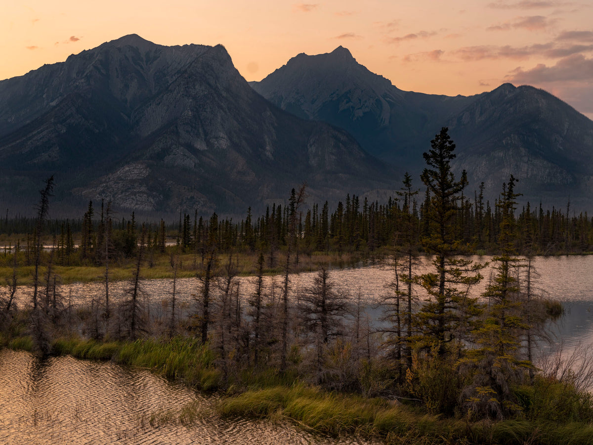 A scenic landscape photograph of rugged mountains rising above a calm lake at sunset, with a line of dark evergreen trees in the foreground and warm orange-pink sky.
